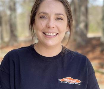 Raegen is wearing a black servpro shirt smiling looking into the camera outside with some trees in the background